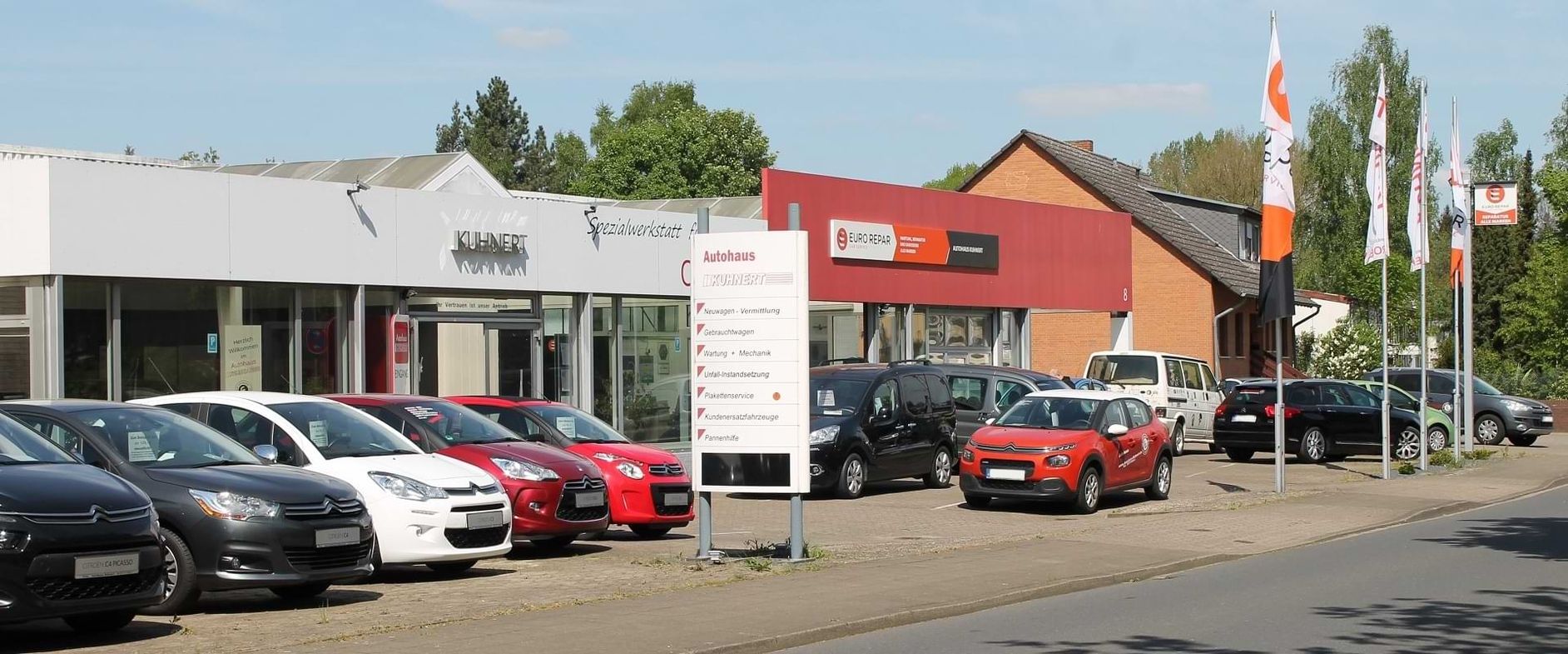 Ein Autohaus mit verschiedenen Fahrzeugen und einem auffälligen Schild, vor einem klaren blauen Himmel und üppigem Grün im Hintergrund.