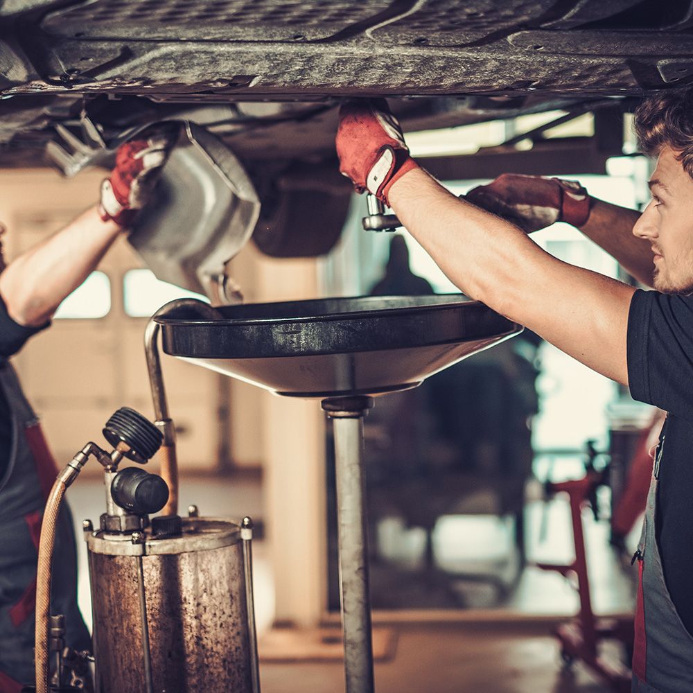 Zwei Mechaniker arbeiten unter einem Auto, einer gießt Öl in eine Auffangwanne, während der andere dabei hilft.